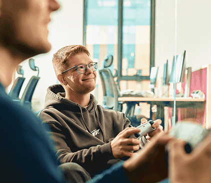 Dwie osoby grające w gry wideo w otoczeniu biurowym, z jedną osobą uśmiechniętą i trzymającą kontroler. Komputery i krzesła są w tle.