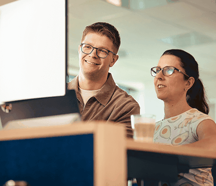 Dwie osoby w okularach uśmiechają się i patrzą na ekran komputera w biurze. Na biurku stoi szklanka wody.
