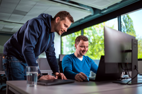 Two men in a bright office, one standing and pointing at a computer screen, the other seated and smiling, with large windows in the background.