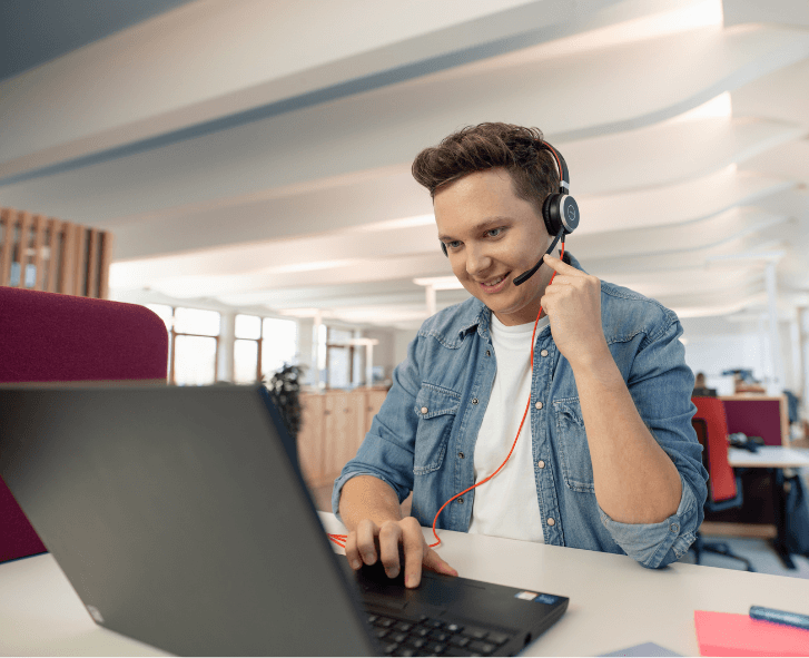 Młody mężczyzna w dżinsowej koszuli, noszący zestaw słuchawkowy, uśmiechnięty podczas pracy na laptopie w nowoczesnym biurze.