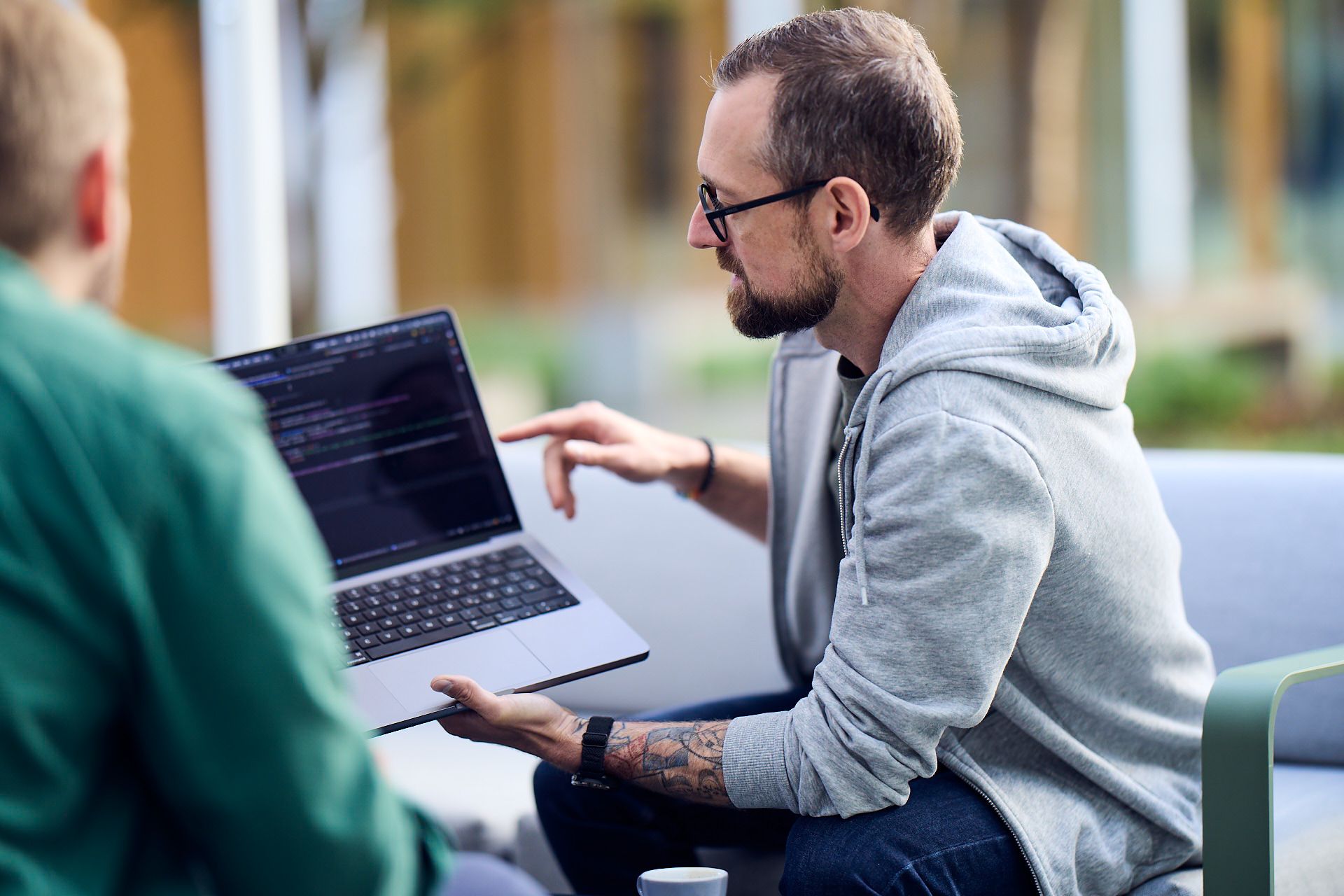 Ein Mann in Kapuzenpulli sitzt draußen und zeigt seinem Kollegen Code auf seinem Laptop
