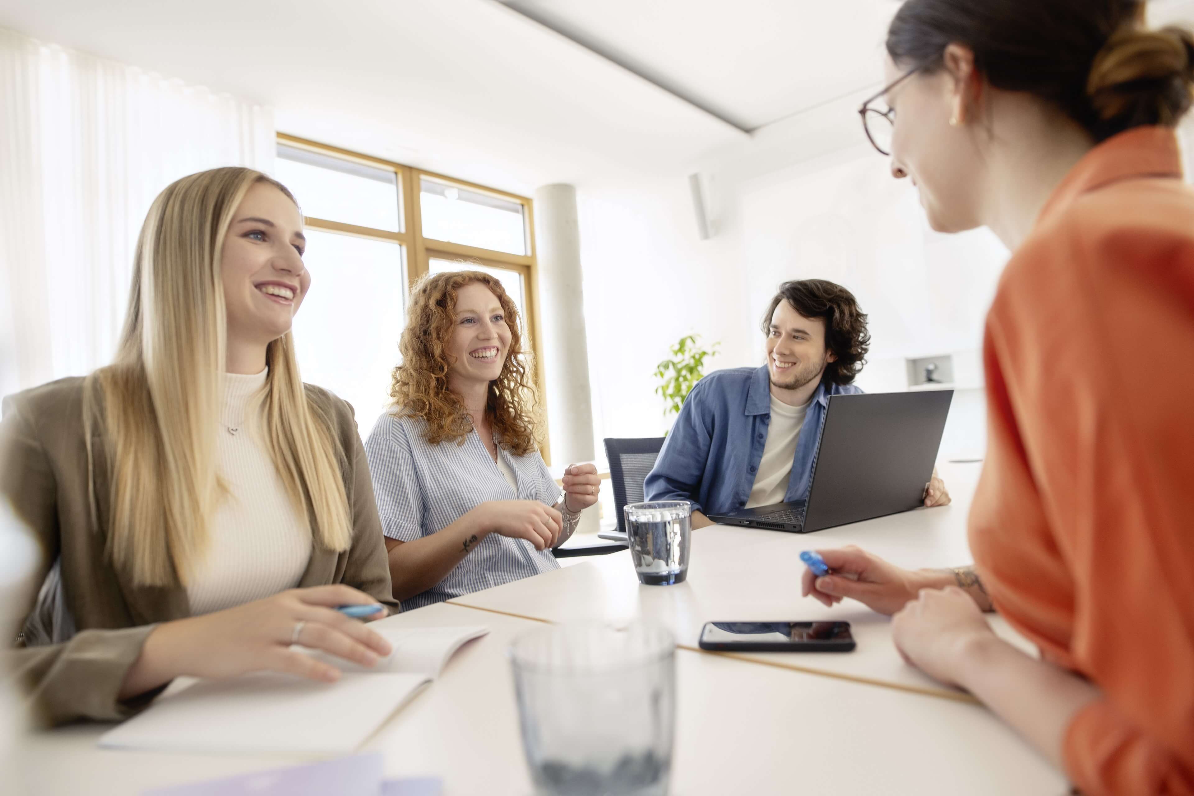Drei junge IT-Mitarbeiterinnen und ein junger IT-Mitarbeiter sitzen in einem Meetingraum im dm-dialogicum und lachen gemeinsam