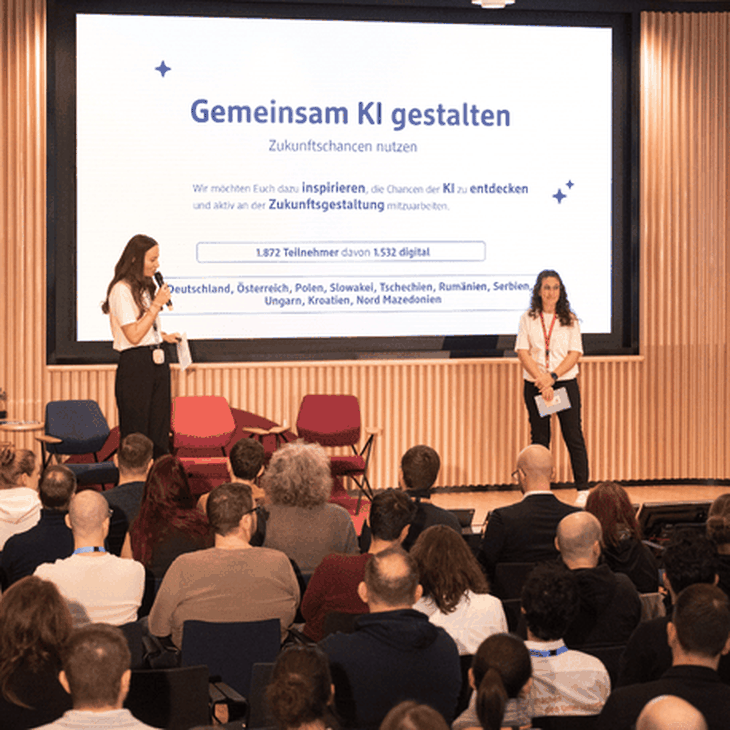 Zwei IT-Kolleginnen präsentieren bei der KI Townhall von dmTECH vor Publikum. Im Hintergrund zeigt ein Bildschirm die Überschrift "Gemeinsam KI gestalten".